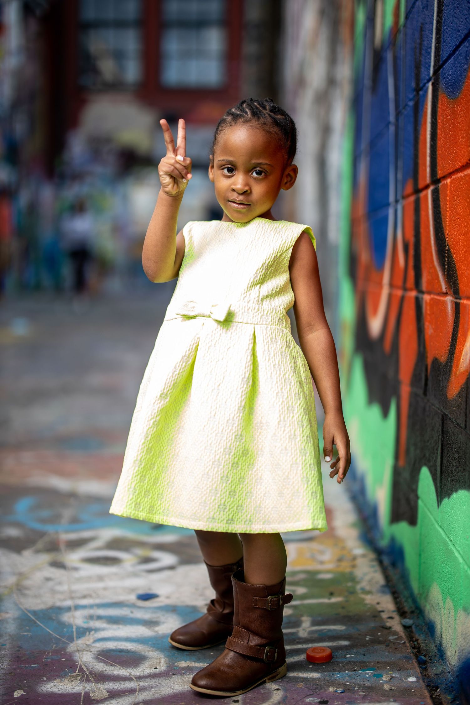 Small child in yellow dress making peace sign against graffiti background.