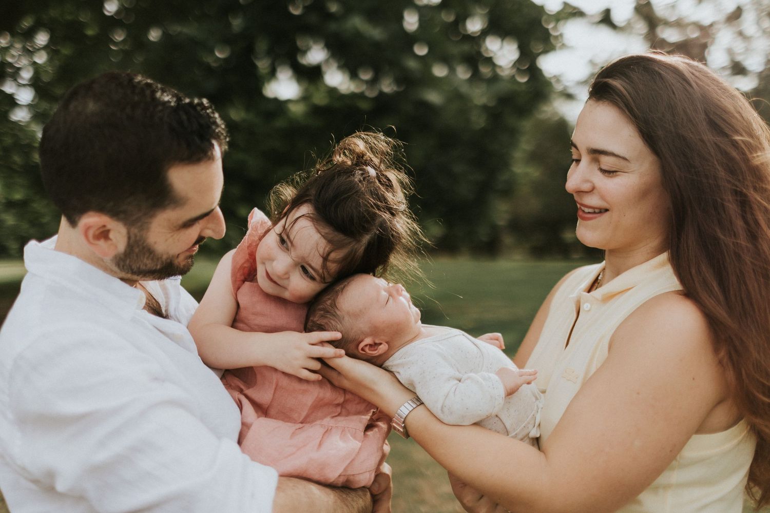 Parents lovingly holding newborn baby while older sibling leans in for a kiss outdoors.