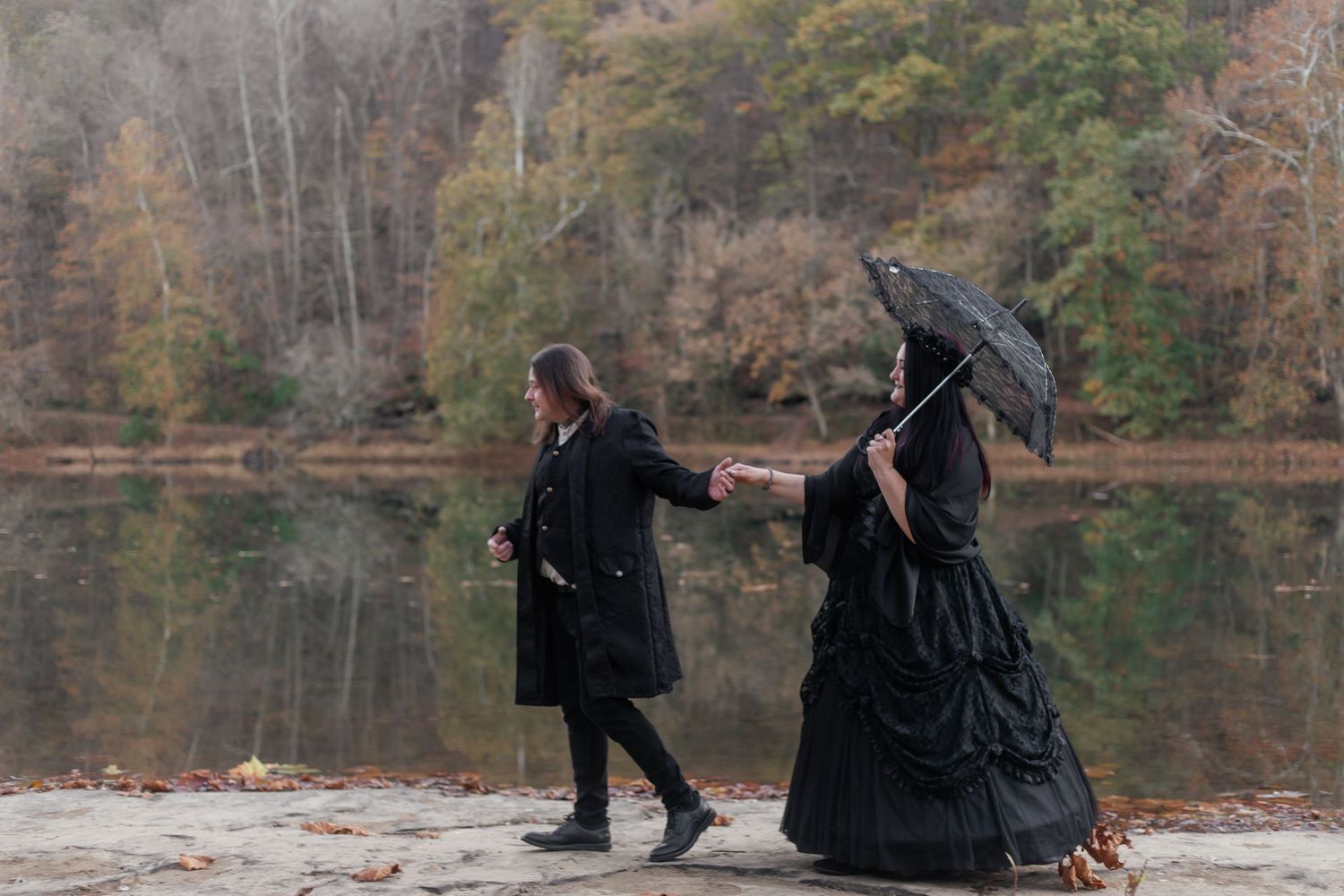 Two people in dark clothing walk along a lakeside path holding hands, one carrying an umbrella.