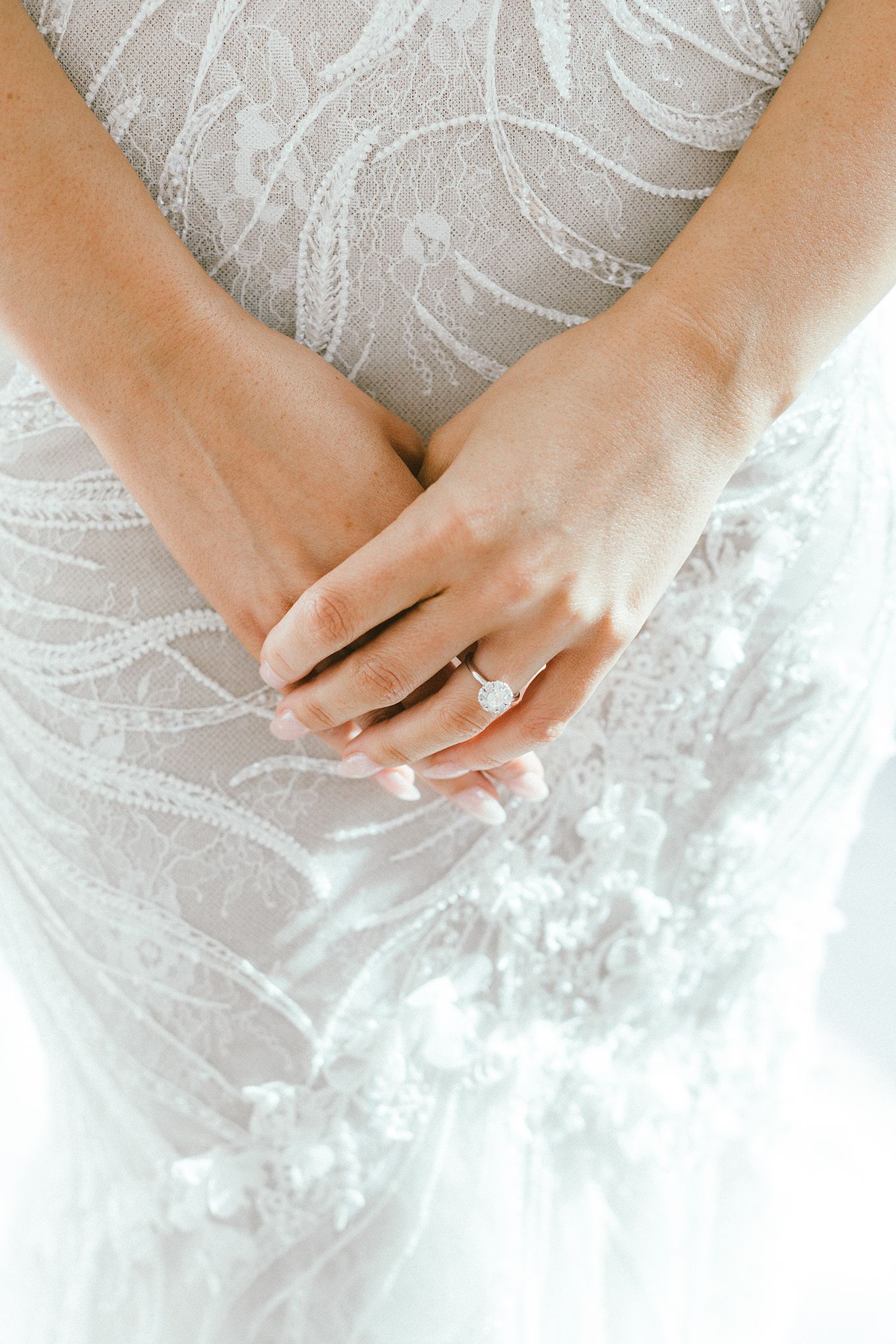 Hands displaying a sparkling engagement ring against a white lace dress.