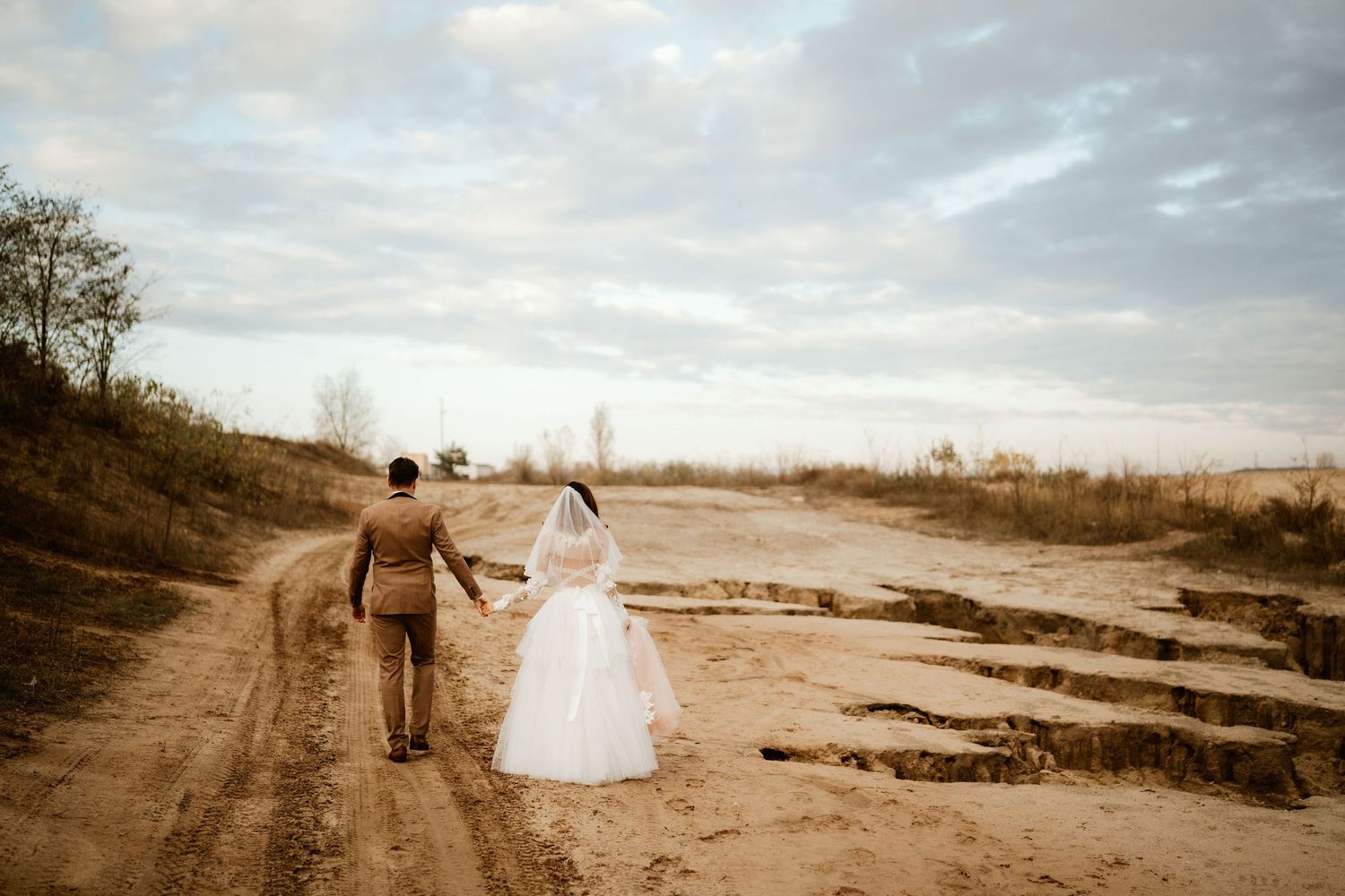 Couple walking along dirt path in wedding attire during sunset.
