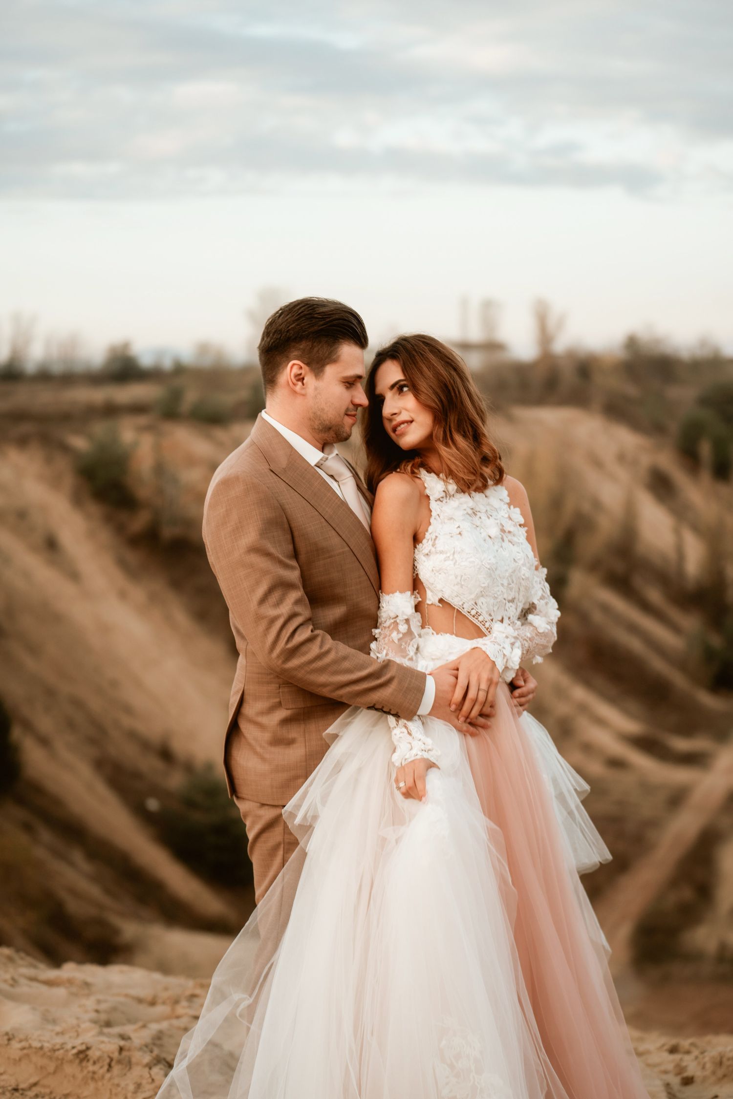 A romantic wedding photo on a desert cliff featuring a brown suit and blush tulle gown at sunset.