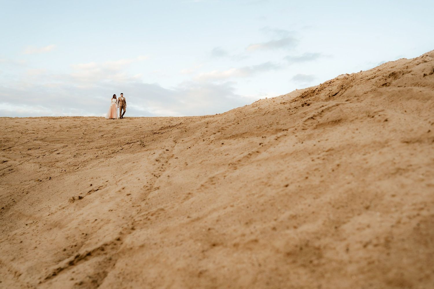 Silhouettes of a couple standing on a sandy hill against cloudy sky during a wedding photoshoot.
