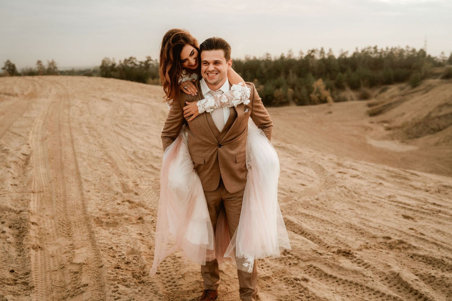 Joyful piggyback wedding photo in desert setting with flowing dress and brown suit.