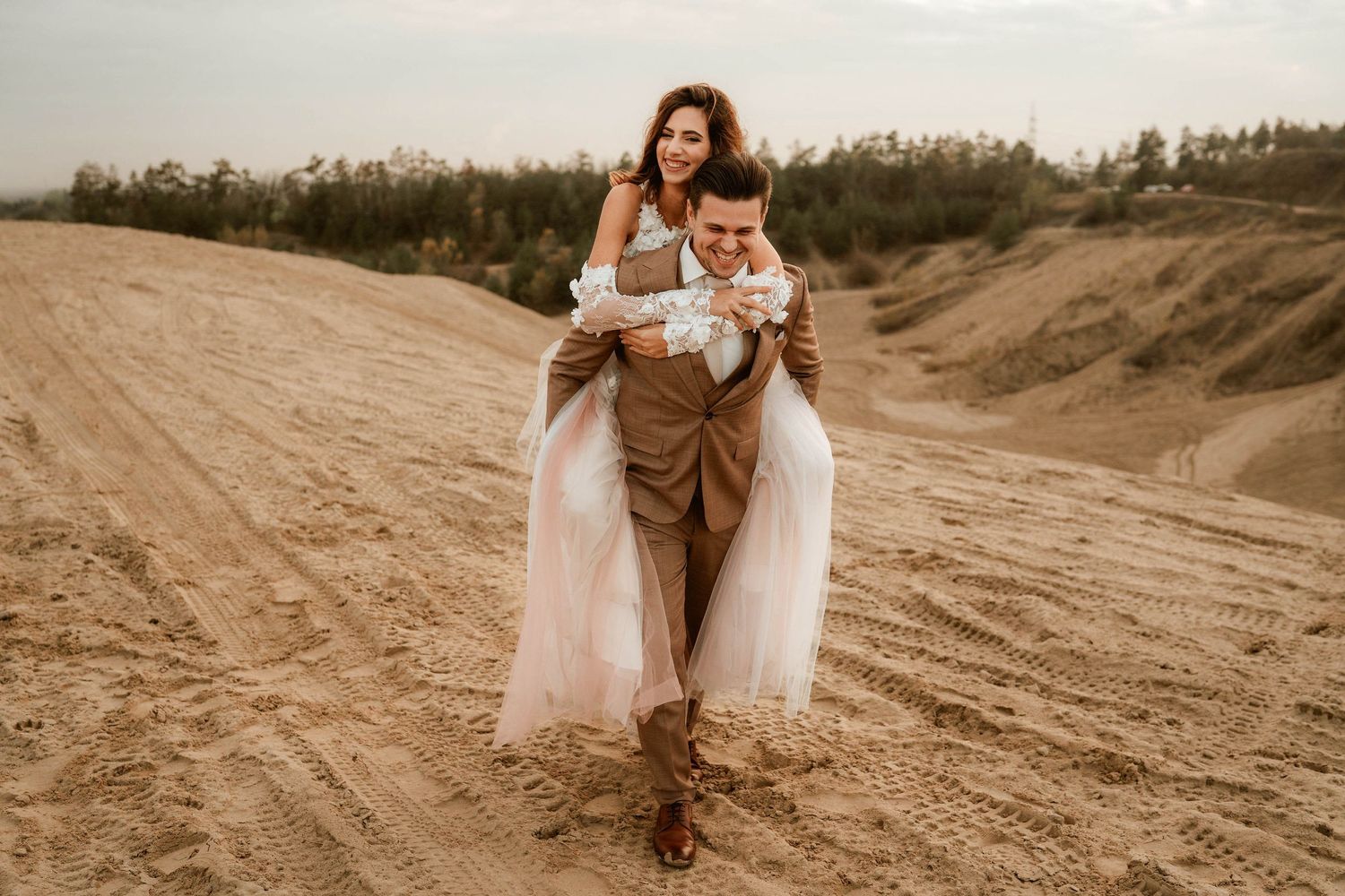 Playful wedding pose on sandy terrain with flowing tulle dress and casual romantic styling.