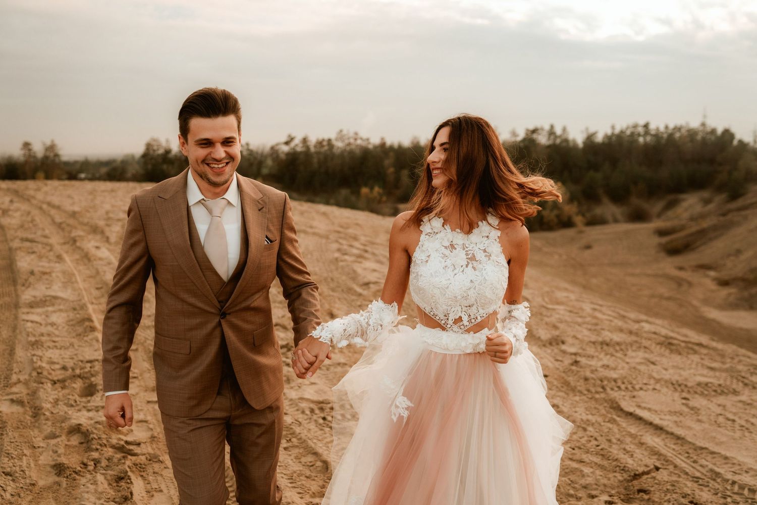 Candid desert wedding photo with couple walking hand in hand in blush gown and brown suit.
