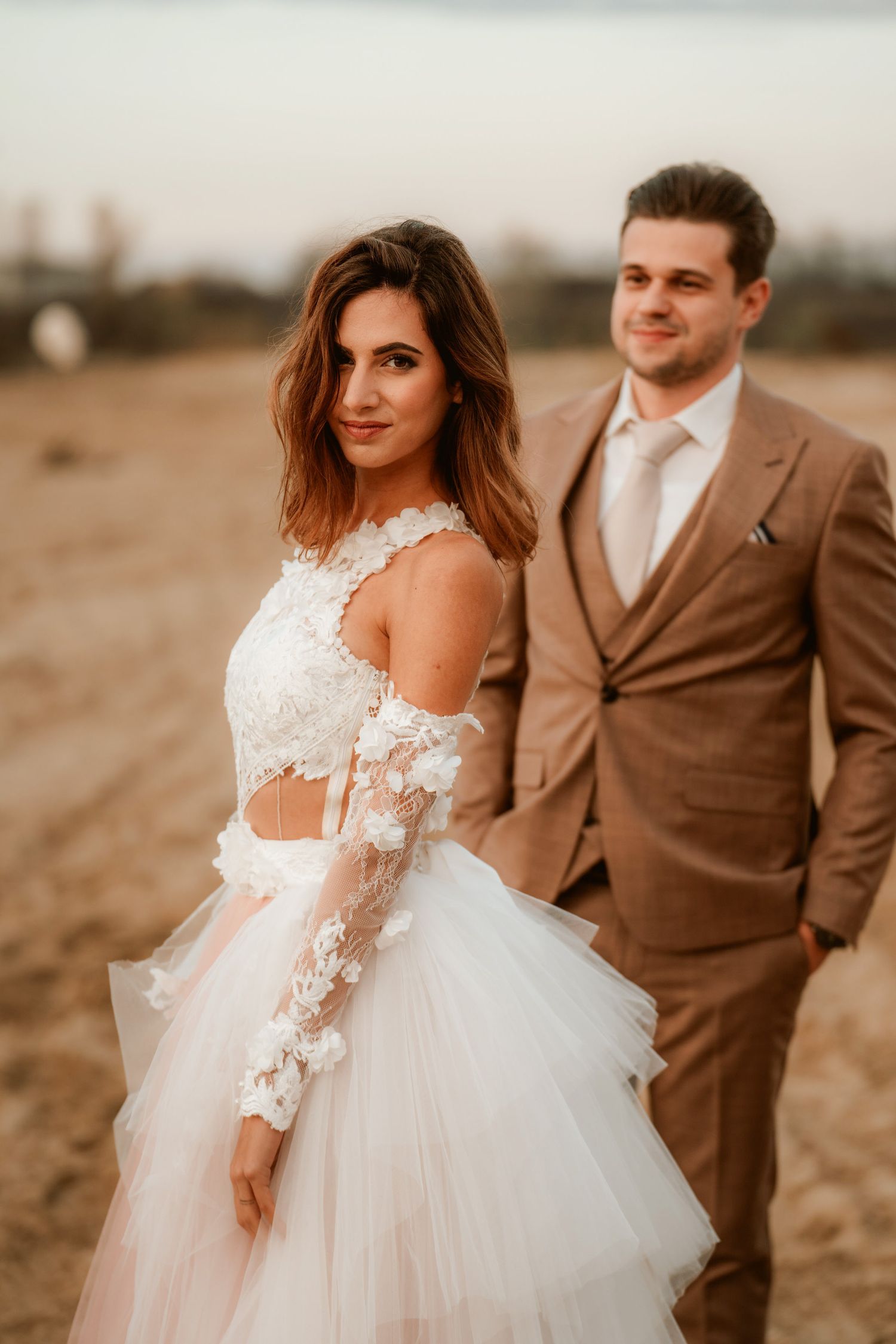 A romantic wedding photo of a couple in formal attire standing on sandy terrain at golden hour.