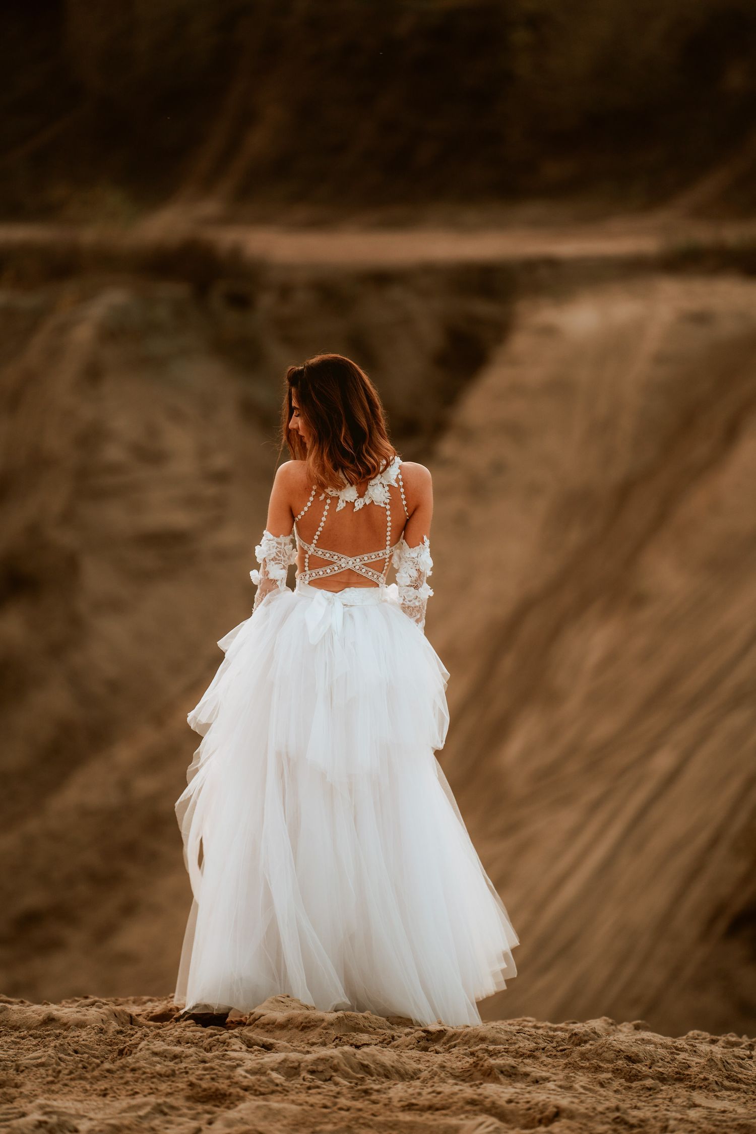 Back view of a bride in a flowing white lace gown with open back design standing on a hill.