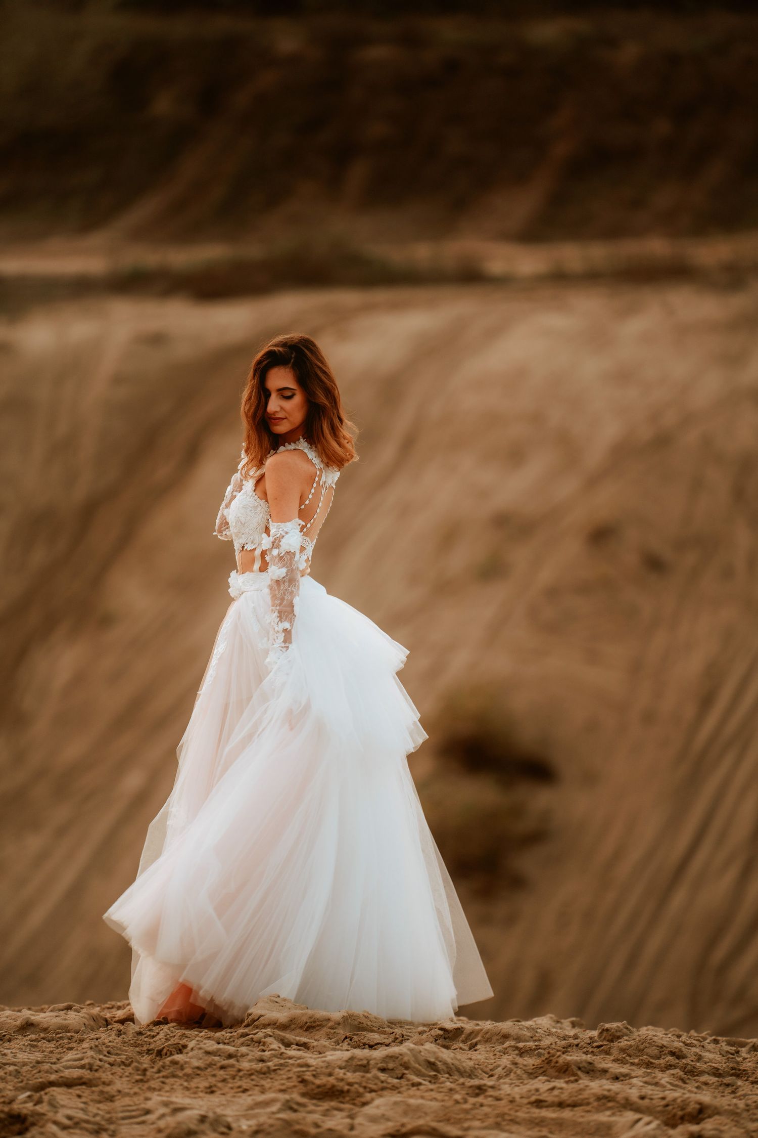 A bride in an elegant white tulle gown poses on a hillside during sunset.