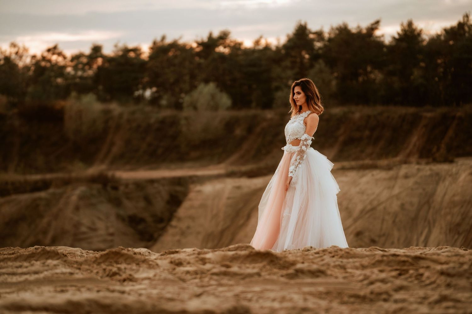 A bride in a flowing white dress stands on a hilltop against a moody sky at dusk.