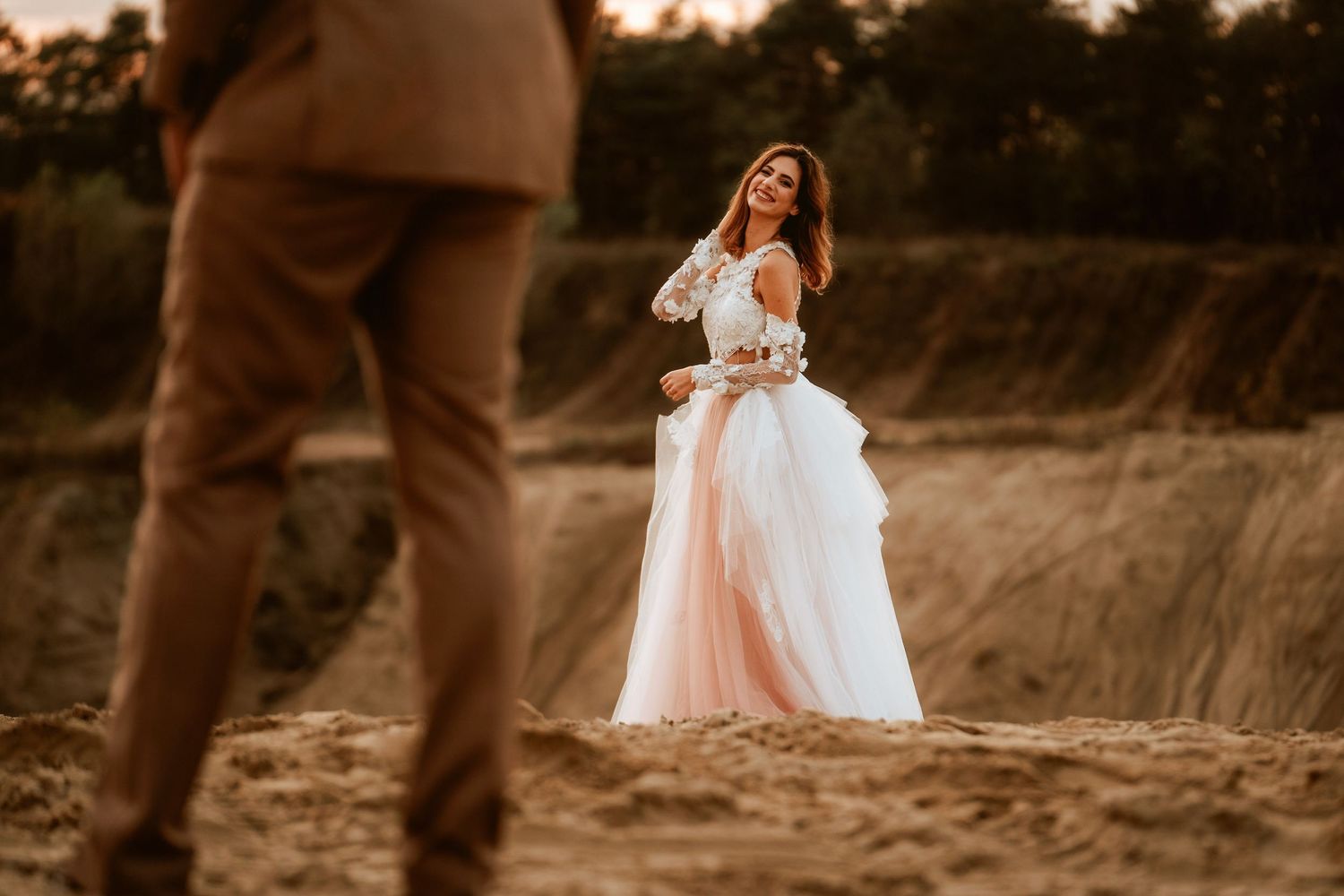 An artistic wedding photo capturing a bride on a hill while groom stands in foreground.