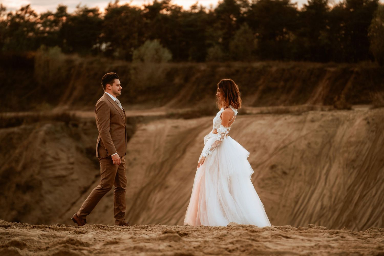 A couple in wedding attire walking on sandy terrain during sunset.