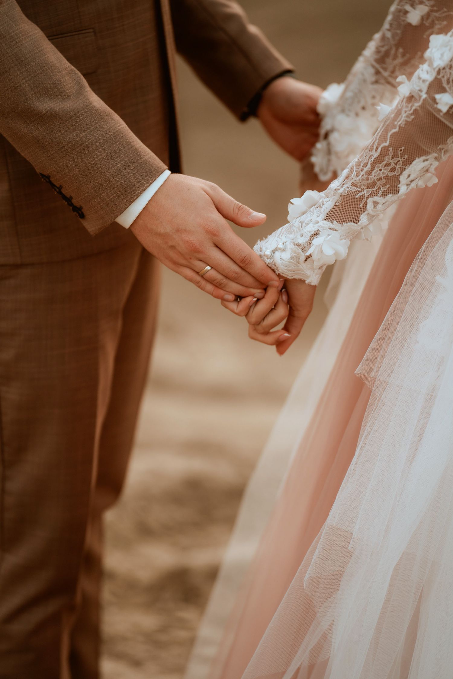 Close-up of newlyweds holding hands, showing details of brown suit and lace wedding dress.