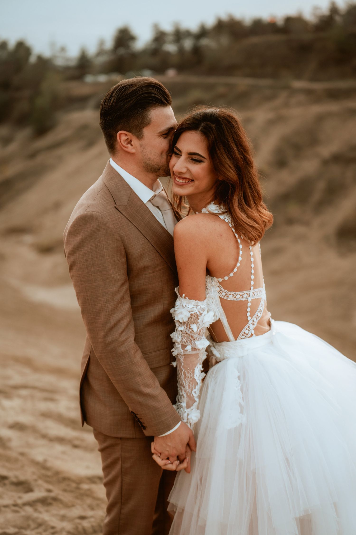 Intimate moment between newlyweds in formal attire on a sandy hillside at sunset.
