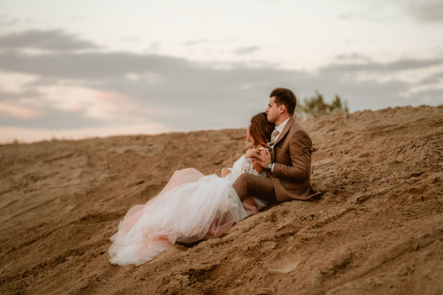 A romantic wedding couple sits together on a sandy hillside during sunset.