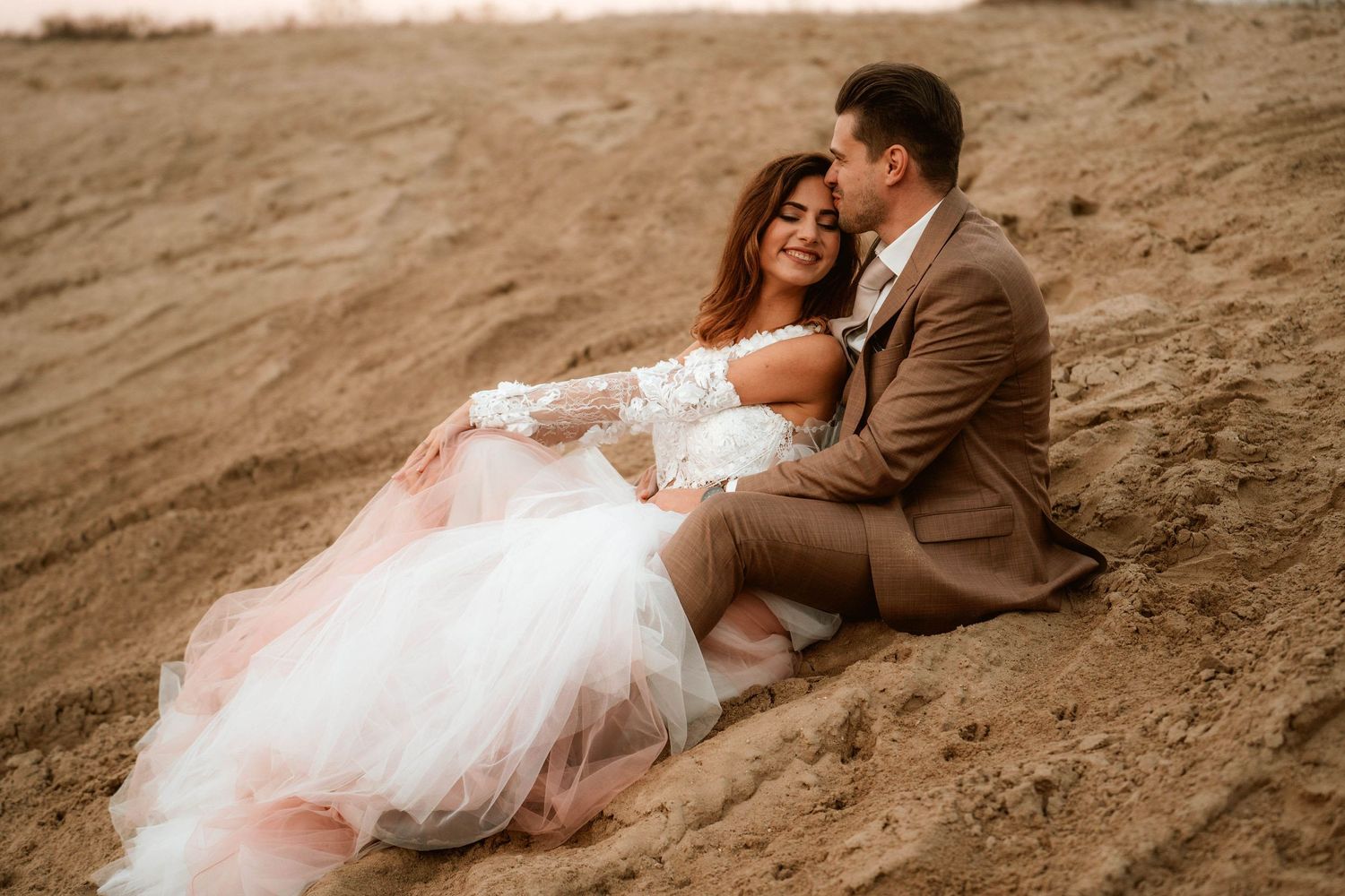 Newlyweds share an intimate moment while sitting on desert sand at dusk.