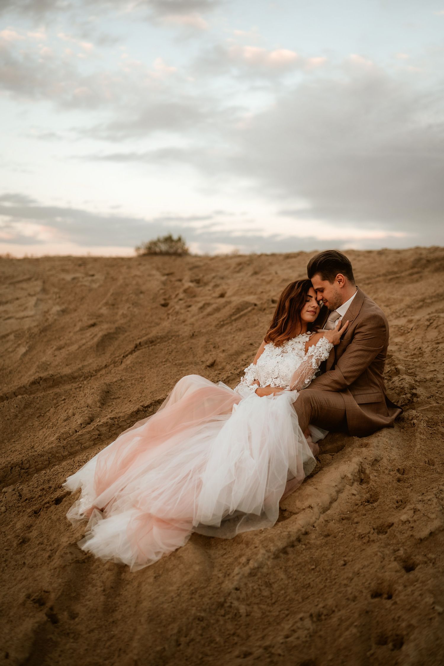 A wedding couple embraces on sandy terrain with cloudy skies in the background.