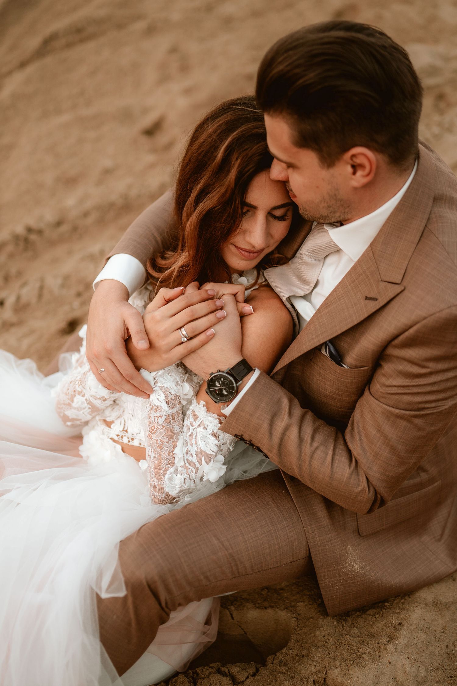 An intimate wedding portrait shows a couple embracing on desert sand.