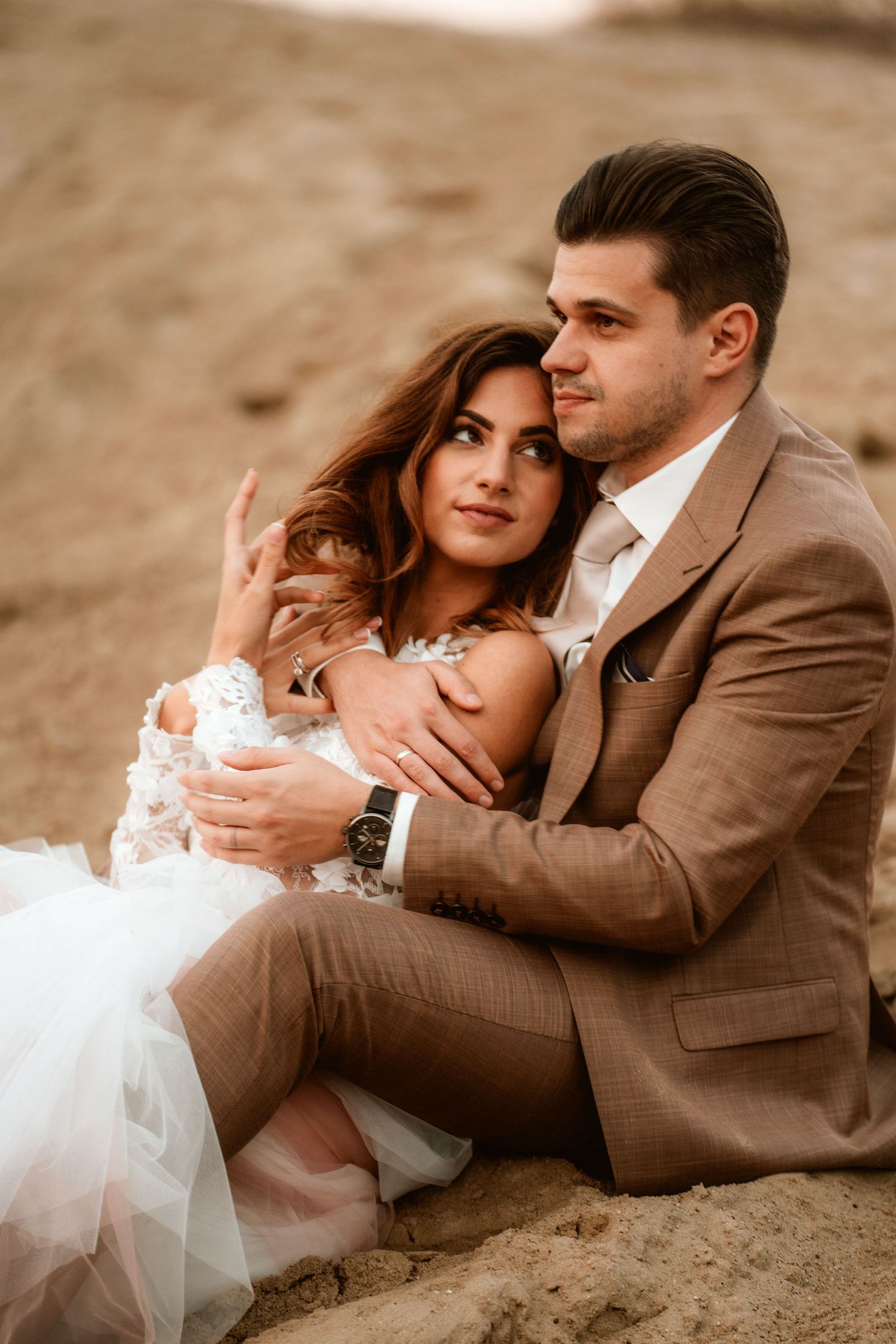 A tender moment between newlyweds in formal attire on sandy terrain.