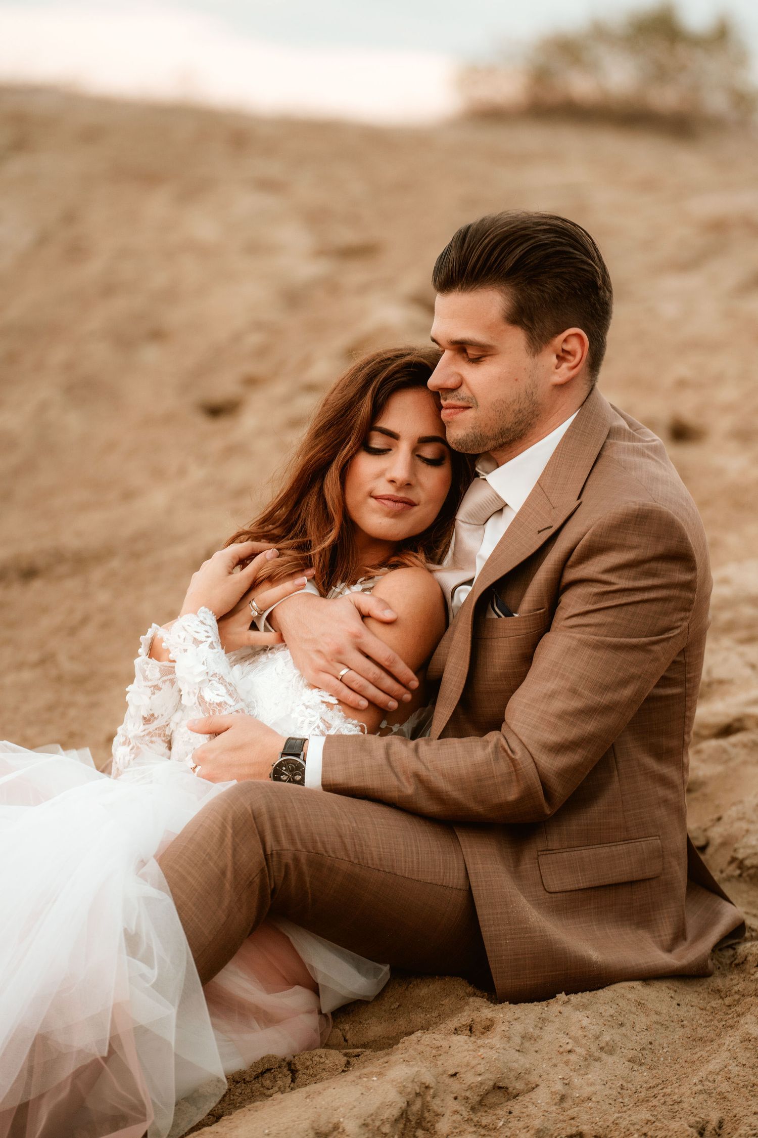 A romantic embrace between newlyweds sitting on sandy ground at sunset.