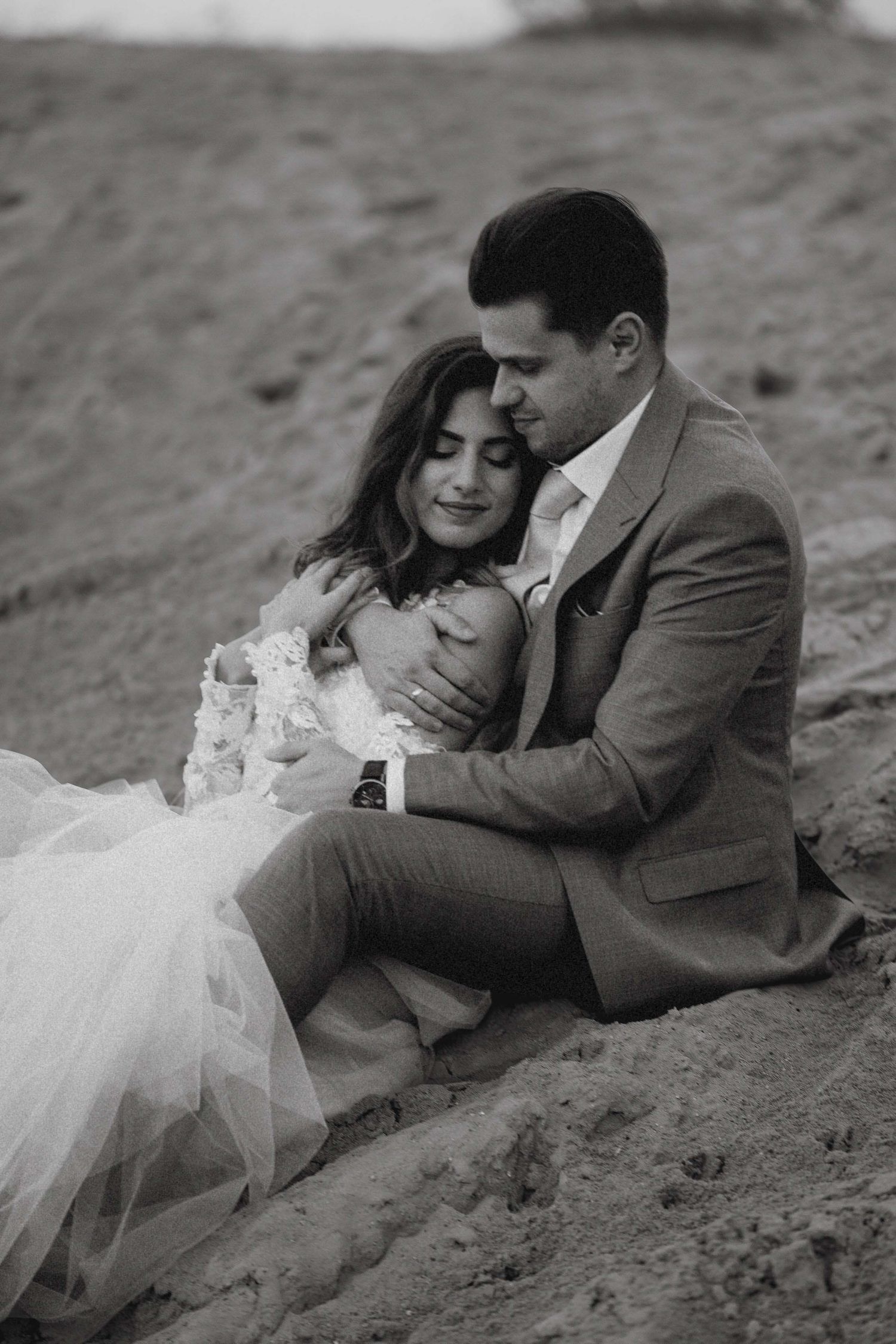 A black and white portrait of a wedding couple sharing a tender moment on sandy terrain.
