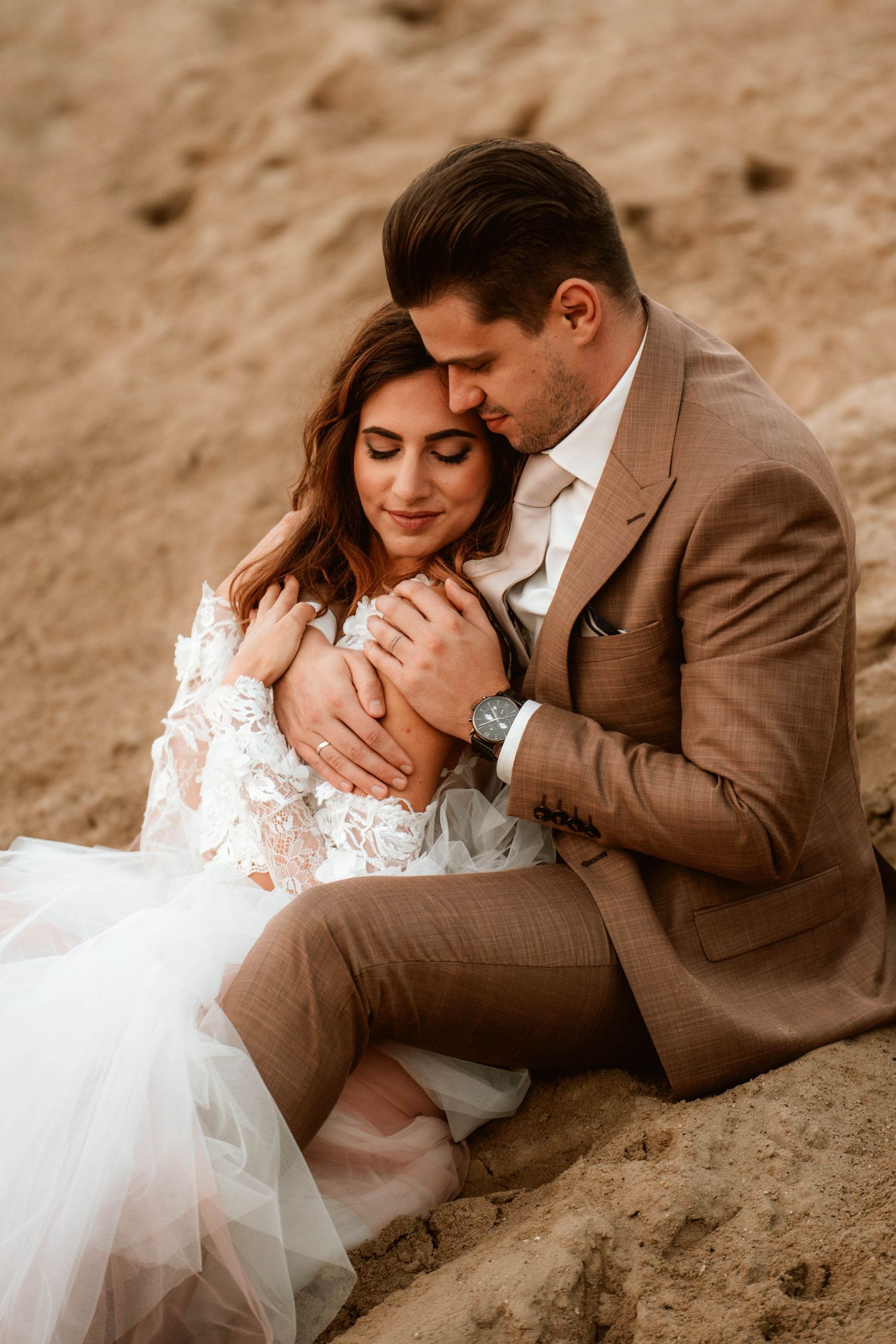 A tender wedding portrait shows couple embracing on sandy terrain at dusk.