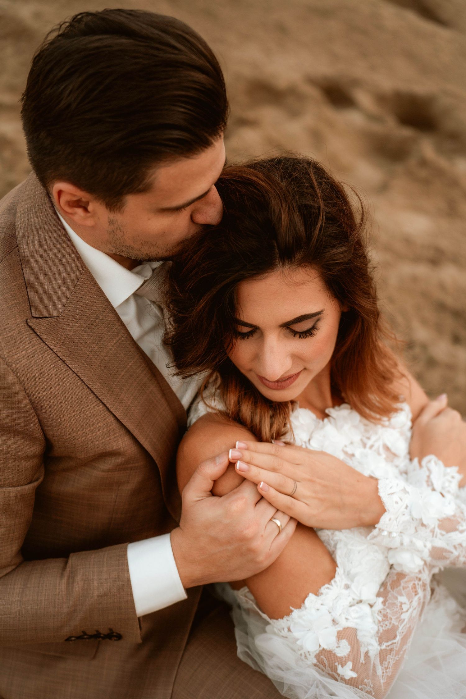 Intimate wedding photo of couple embracing on sandy terrain during golden hour in romantic pose.