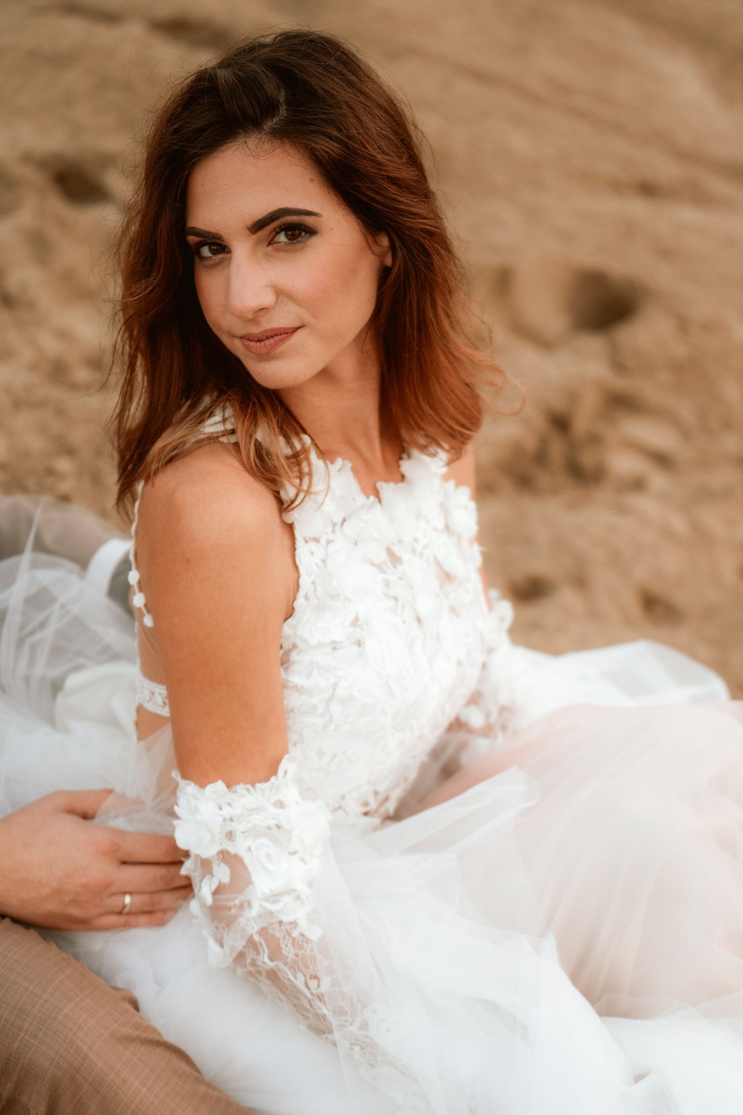 Elegant bride in lace white dress poses against sandy backdrop during outdoor photoshoot.