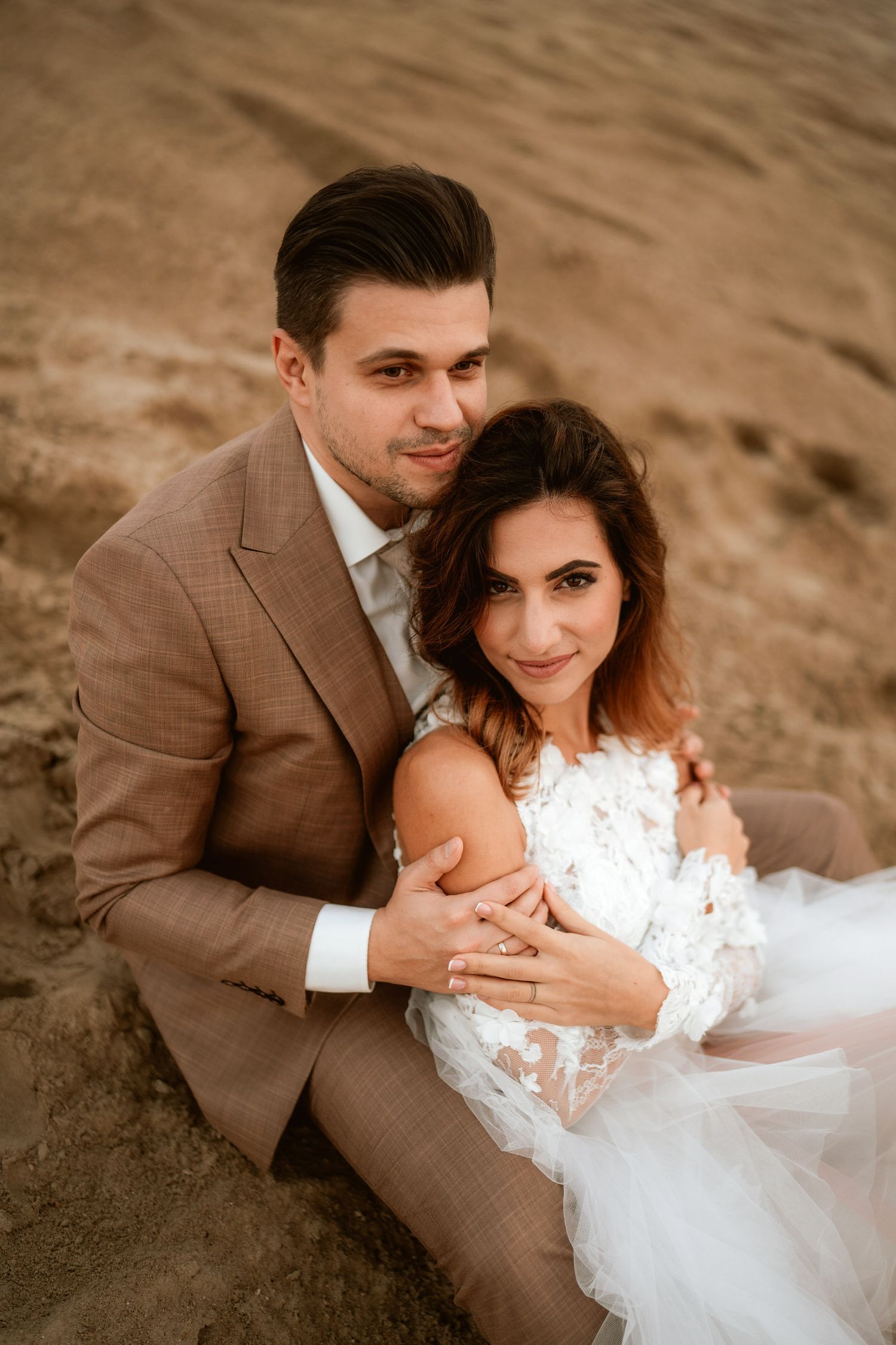 Romantic wedding portrait of couple in brown suit and white dress against desert landscape.