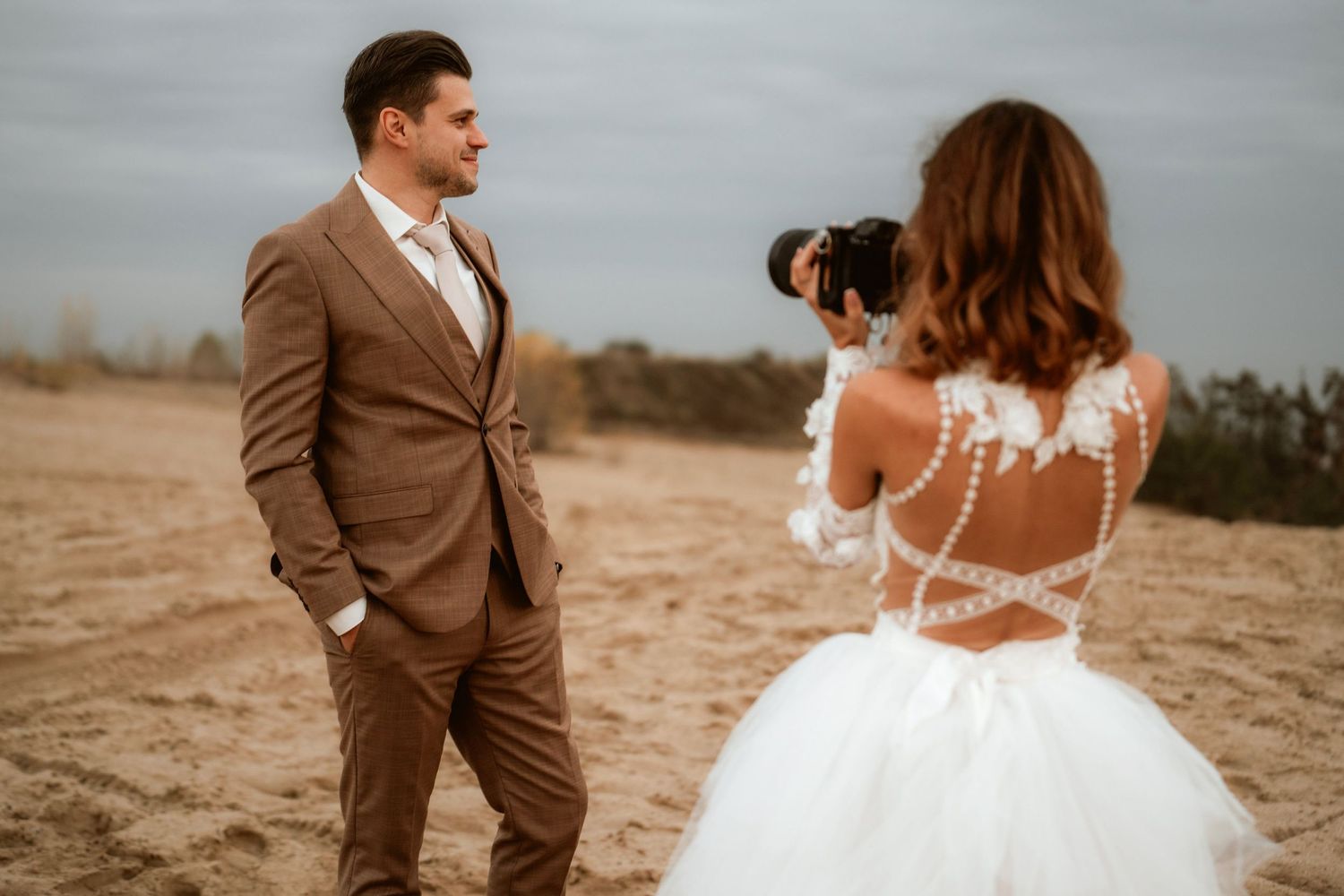 Photographer captures a romantic moment in a tan suit and white wedding dress against a desert landscape.