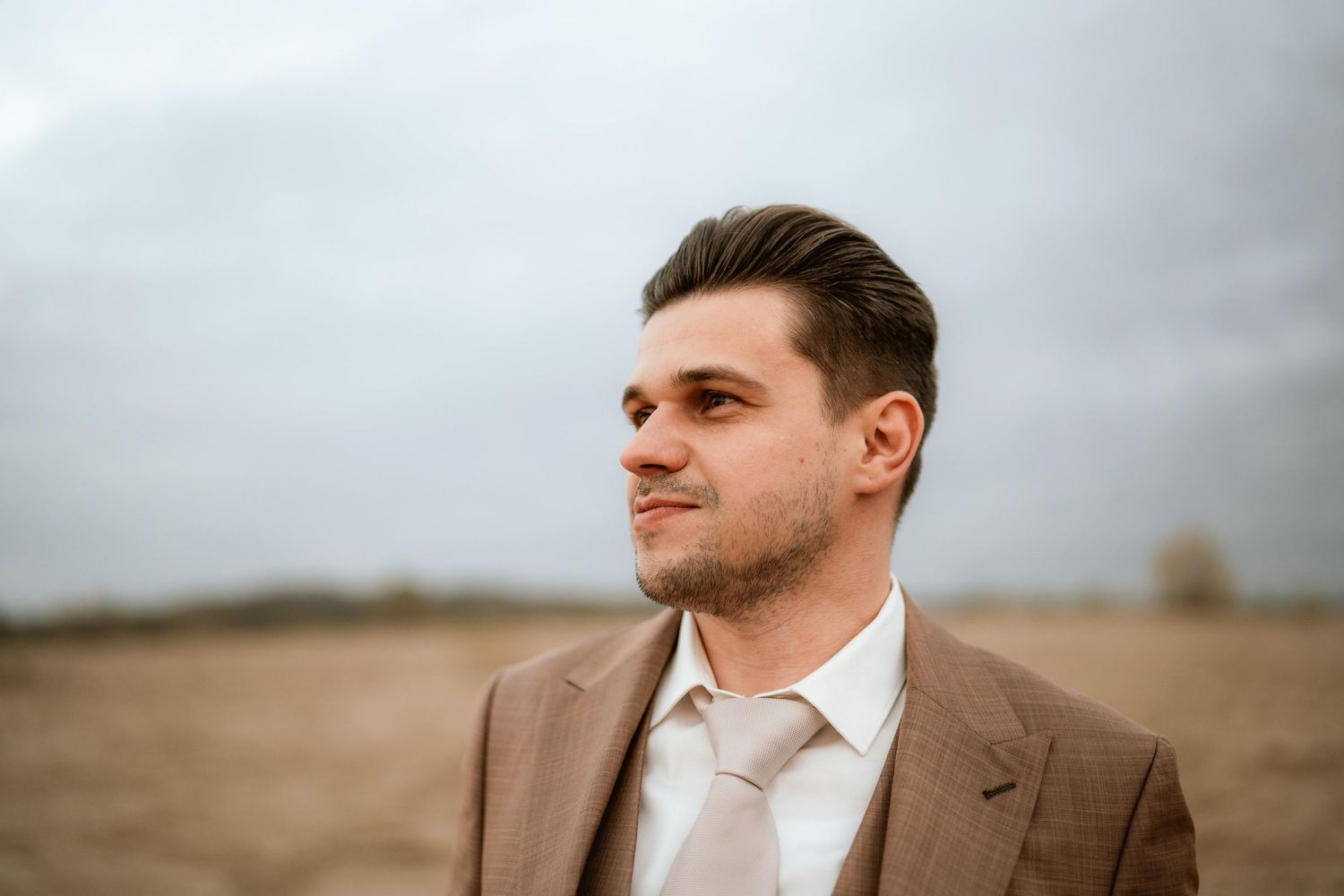 Person in a brown suit with white shirt gazes into the distance on an overcast day.