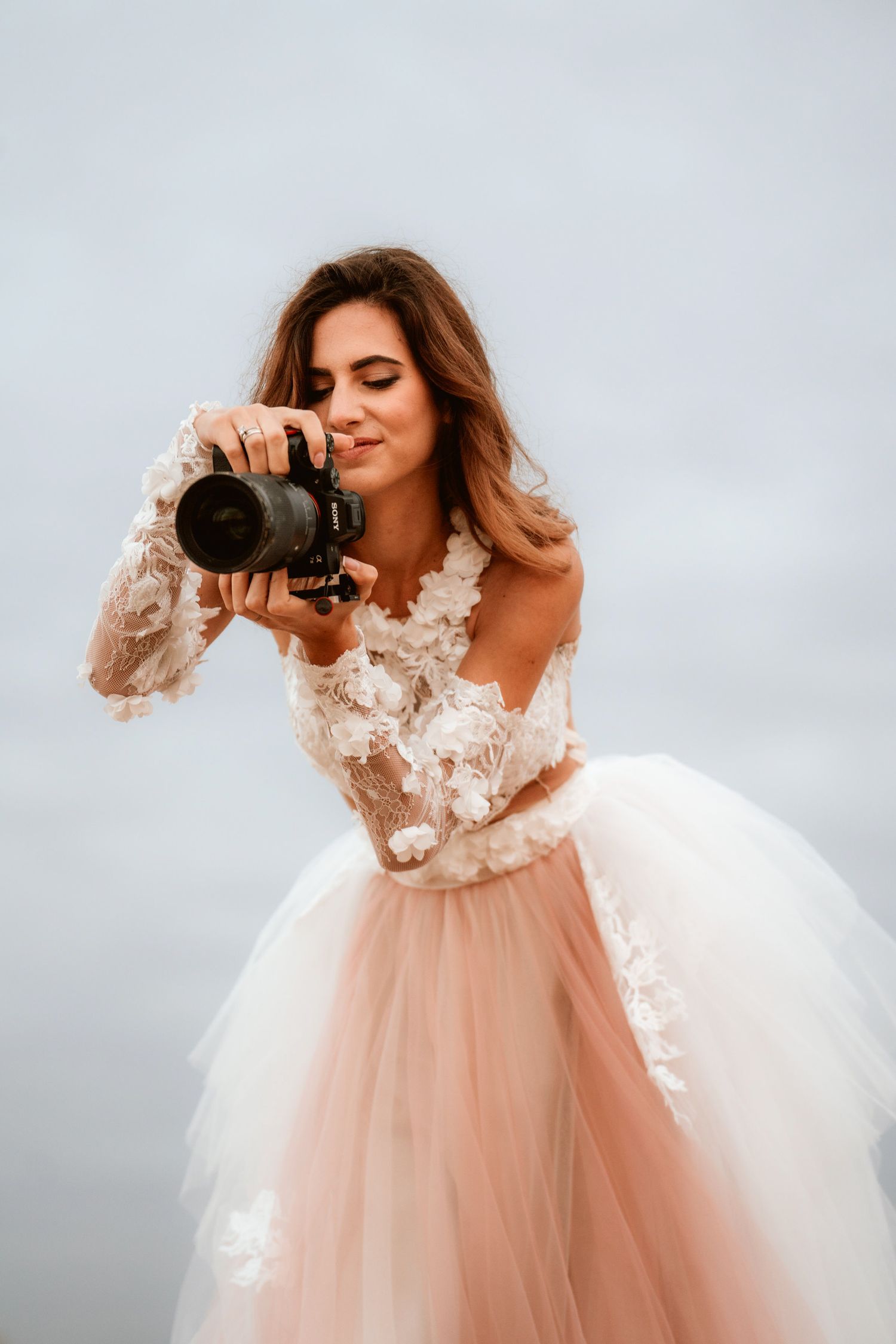 Figure in a blush tulle wedding dress takes photos with a professional camera against cloudy skies.