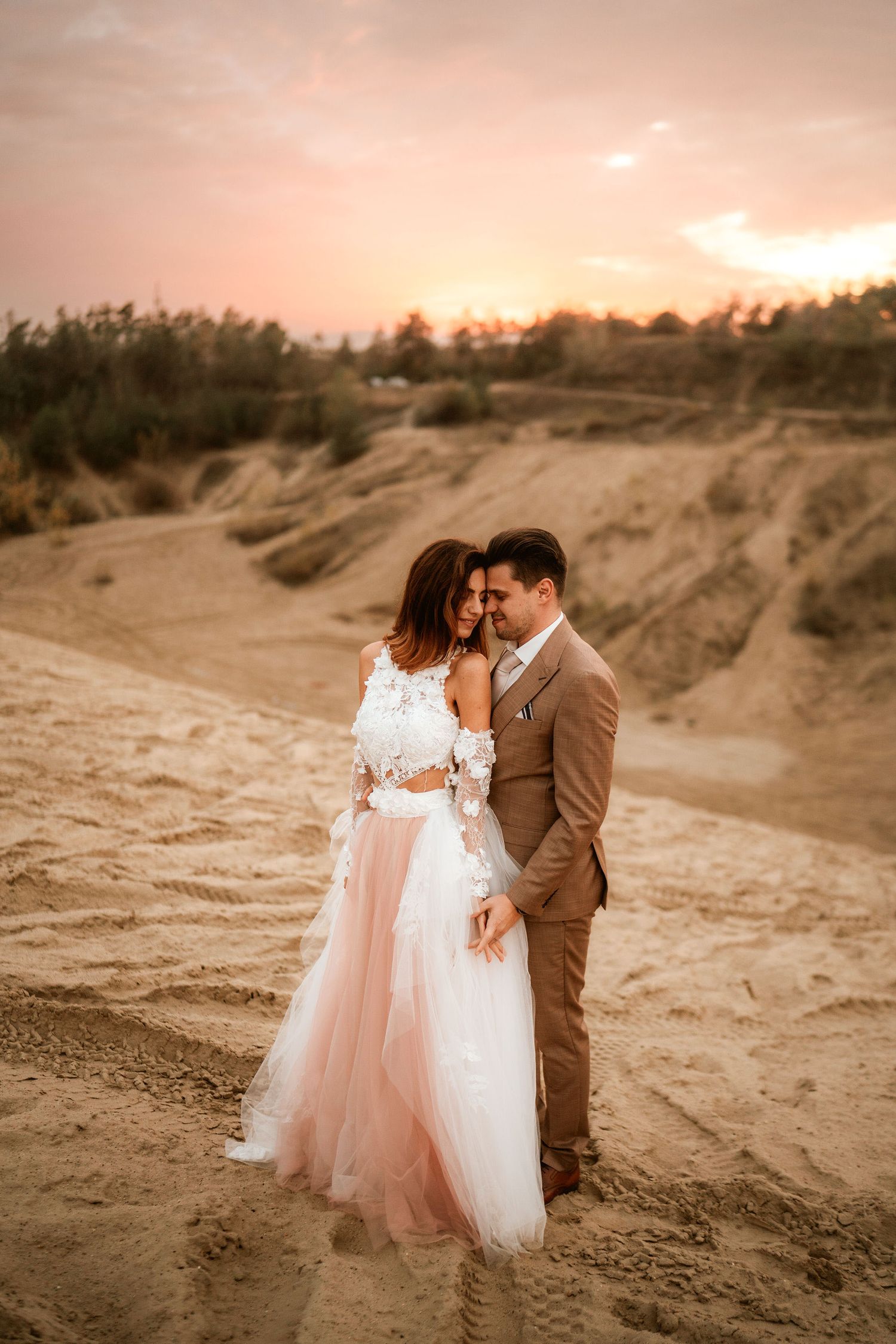 Couple in formal attire poses against sunset-lit desert landscape.