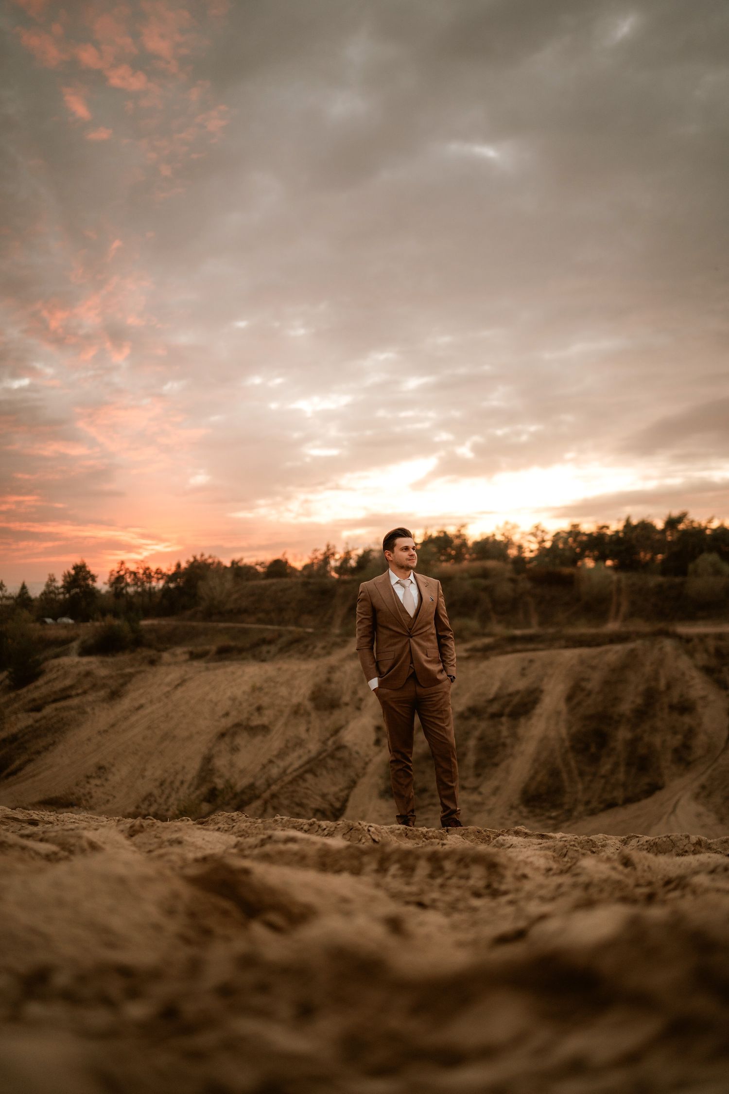 Person in brown suit stands alone against dramatic sunset sky in desert setting.