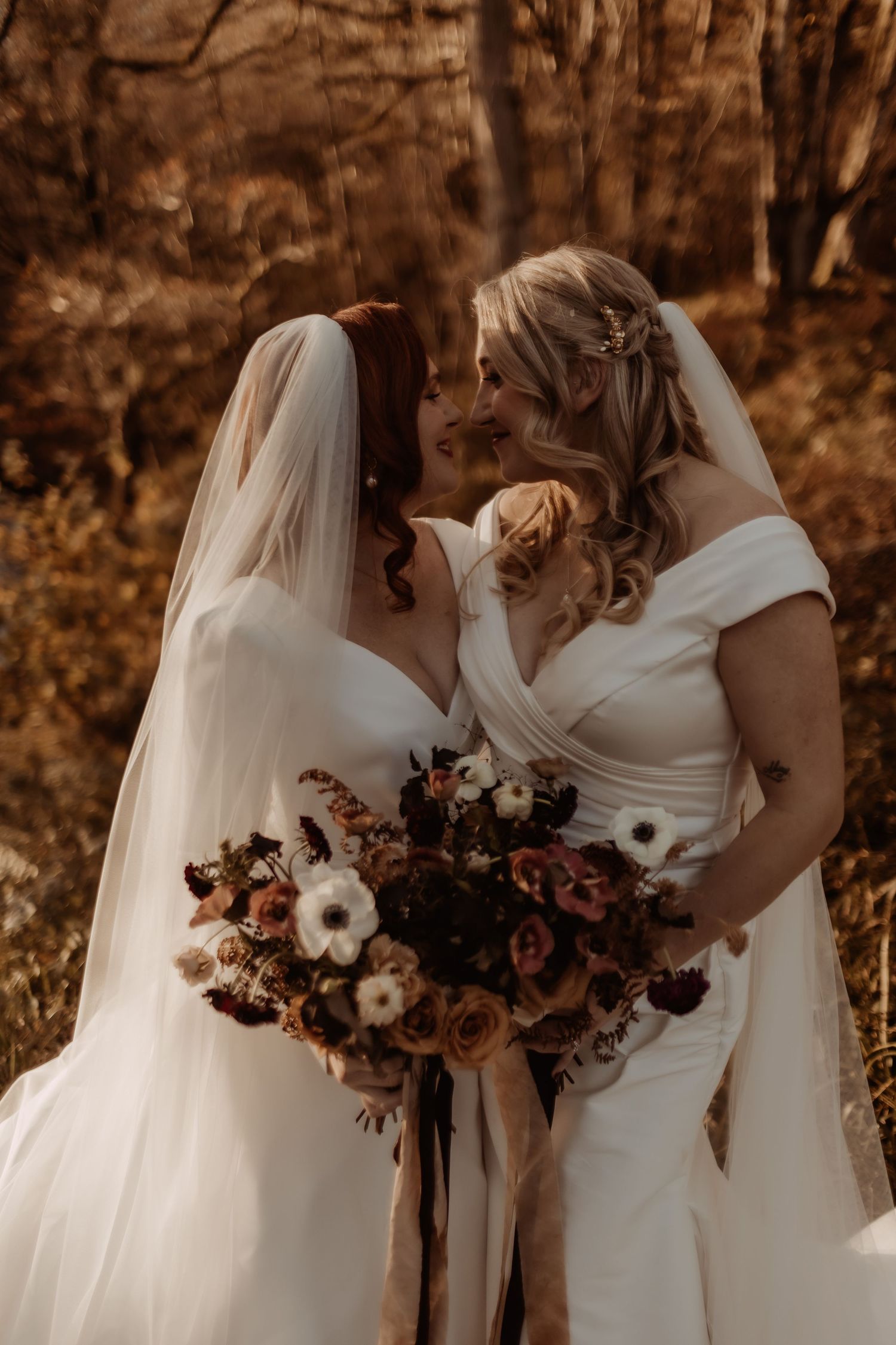 Two brides in white dresses share intimate moment holding autumn-colored wedding bouquets.