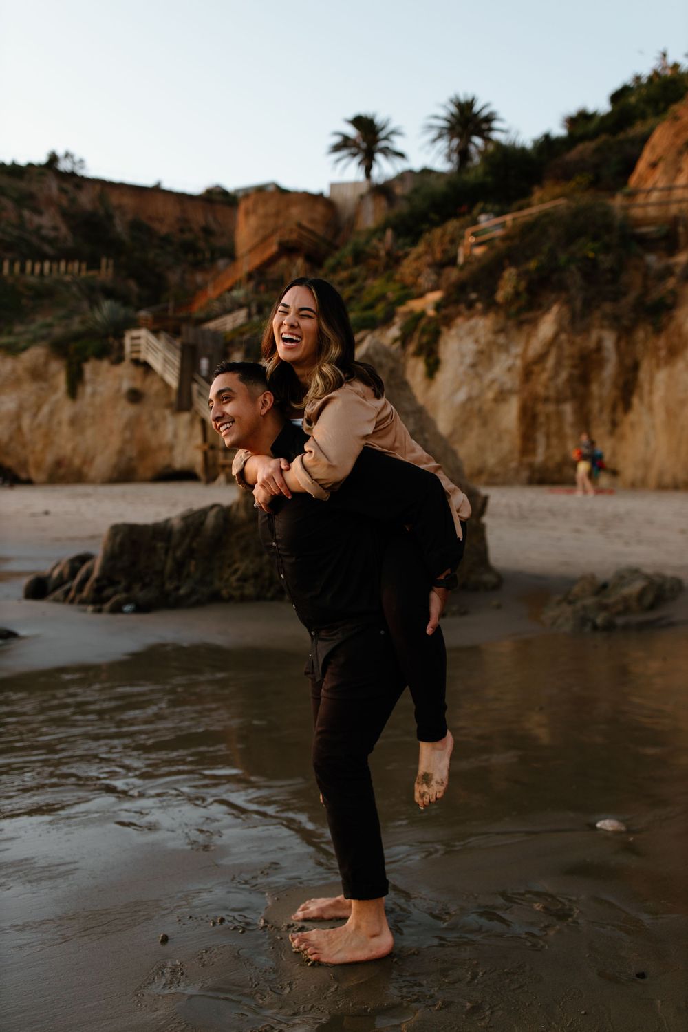 A playful piggyback ride moment captured on a scenic beach with cliffs in the background.
