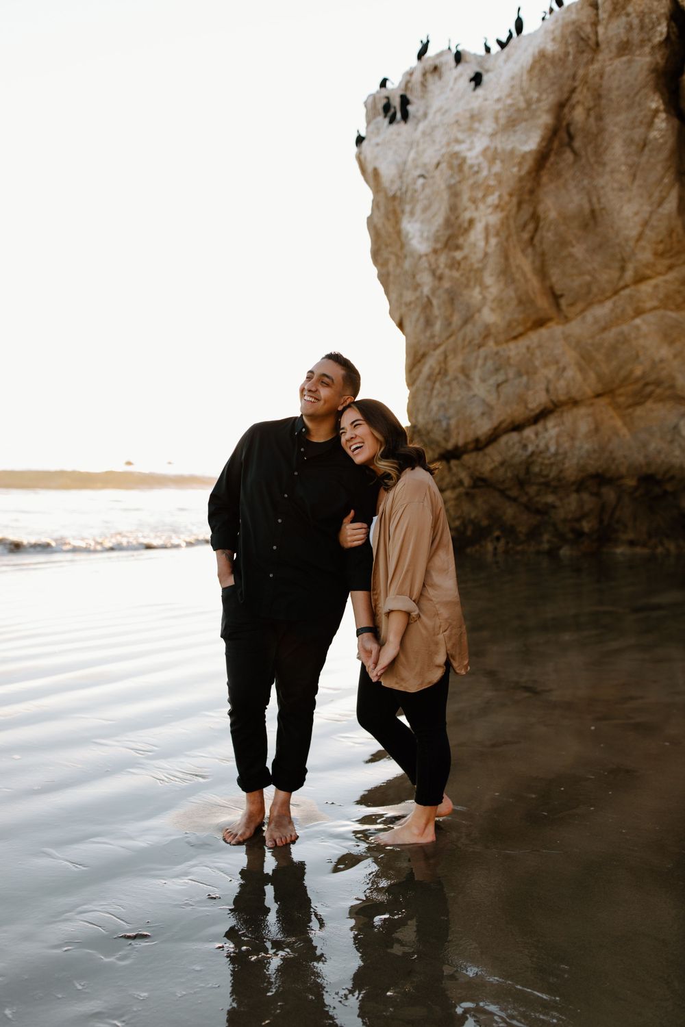 A couple stands barefoot in shallow water against a dramatic coastal cliff backdrop.