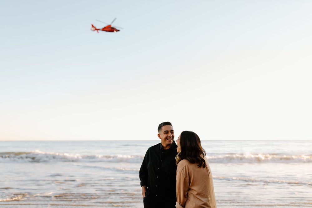 Couple walking along the beach with a helicopter flying overhead and sunset in the distance.
