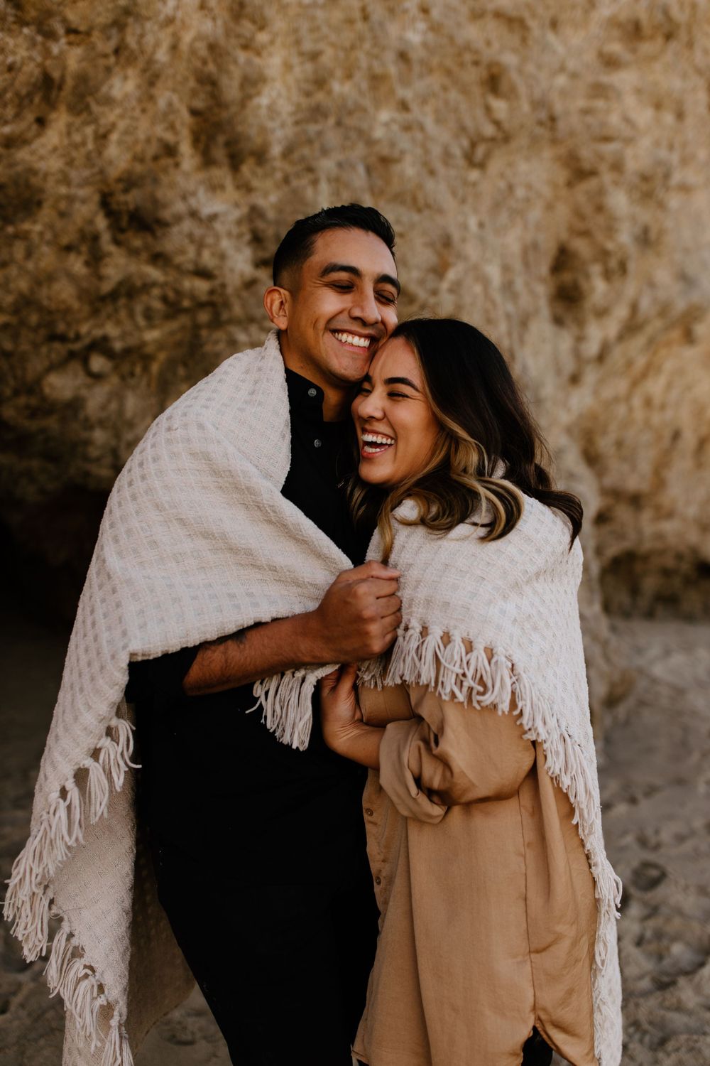 A couple shares laughter while wrapped in a white blanket against a rocky backdrop.