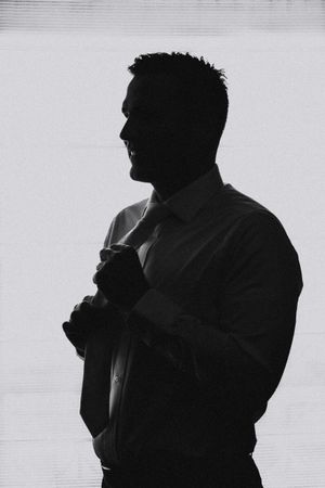 Dramatic black and white silhouette of a person adjusting their tie against a light background.