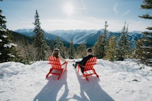 Two red Adirondack chairs overlooking snowy mountain vistas on a sunny day.