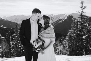 Wedding couple embracing on snowy mountaintop with dramatic mountain backdrop.