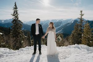 A winter wedding couple stands together on a snowy mountain overlook with evergreen trees.