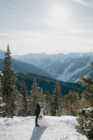 Newlyweds embrace on a snowy mountain ridge surrounded by evergreen trees and dramatic peaks.