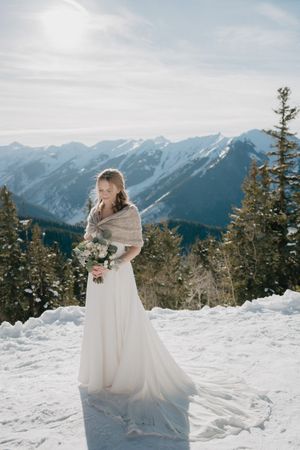 A bride in a flowing white dress and shawl holds a bouquet against a snowy mountain landscape.