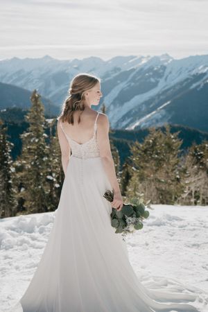 A bride in a flowing white dress stands against dramatic mountain peaks in winter.