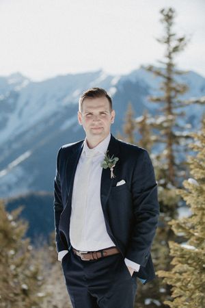 A groom in a navy suit poses against snow-capped mountains and evergreen trees.