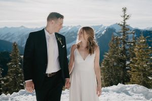 A couple in formal wedding attire stands together on a snowy mountain peak surrounded by evergreen trees and distant peaks.