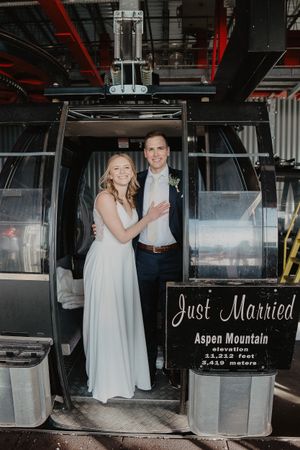 A newlywed couple poses in formal attire at the entrance of an Aspen Mountain gondola with a Just Married sign.
