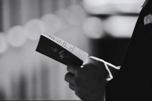 Black and white image of someone reading from a book against a blurred background.